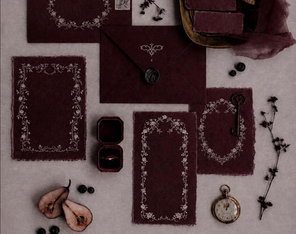 Styled burgundy wedding stationery mockup featuring delicate ivory floral border designs, envelopes, a ring box, and antique-inspired accessories.