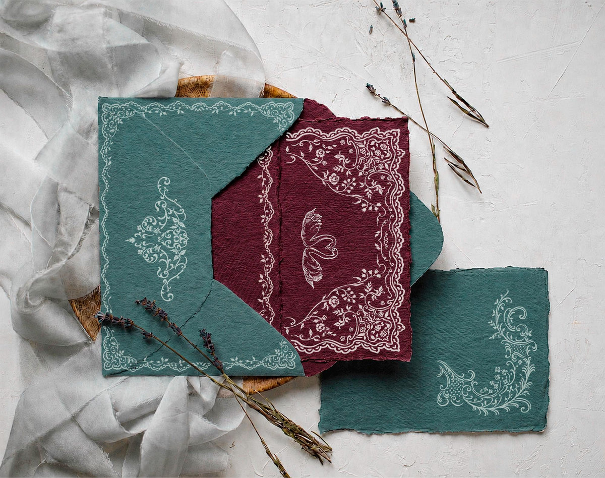 Styled wedding stationery mockup featuring Ivory Lace white ornamental borders on green and burgundy handmade paper.